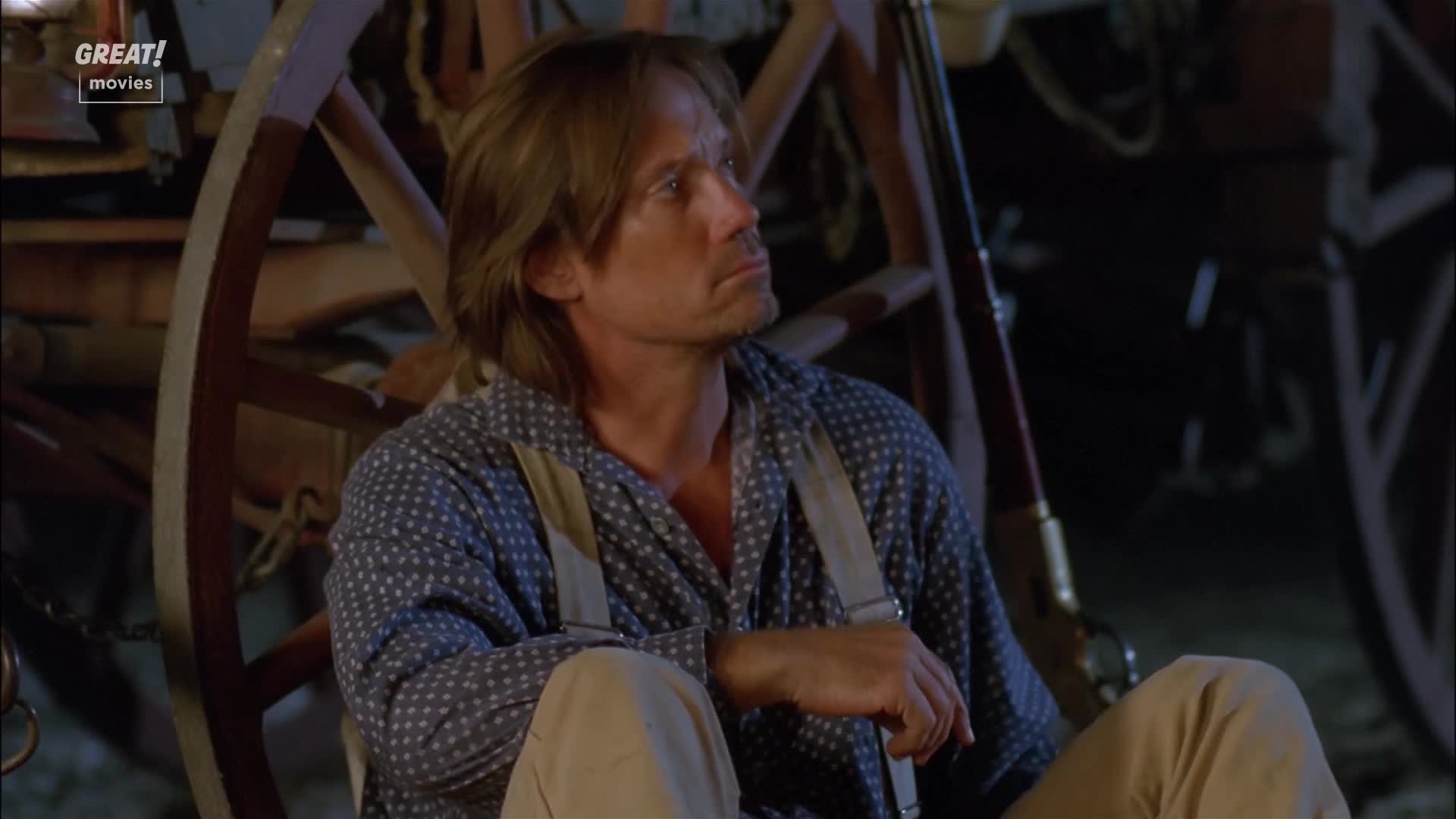 He sits, looking off to the side, lost in thought. The man is dressed in a blue patterned shirt with suspenders, a rifle resting nearby.
