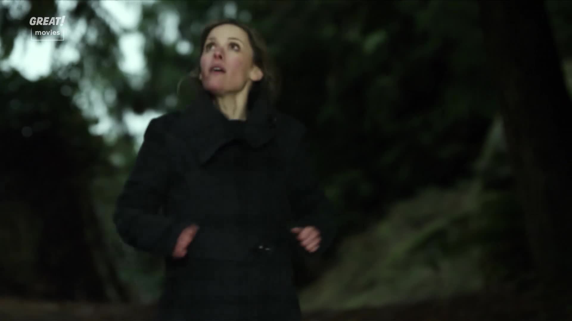 The woman is running, her face upturned with a look of worry.  She's wearing a dark coat, moving quickly through a wooded area.
