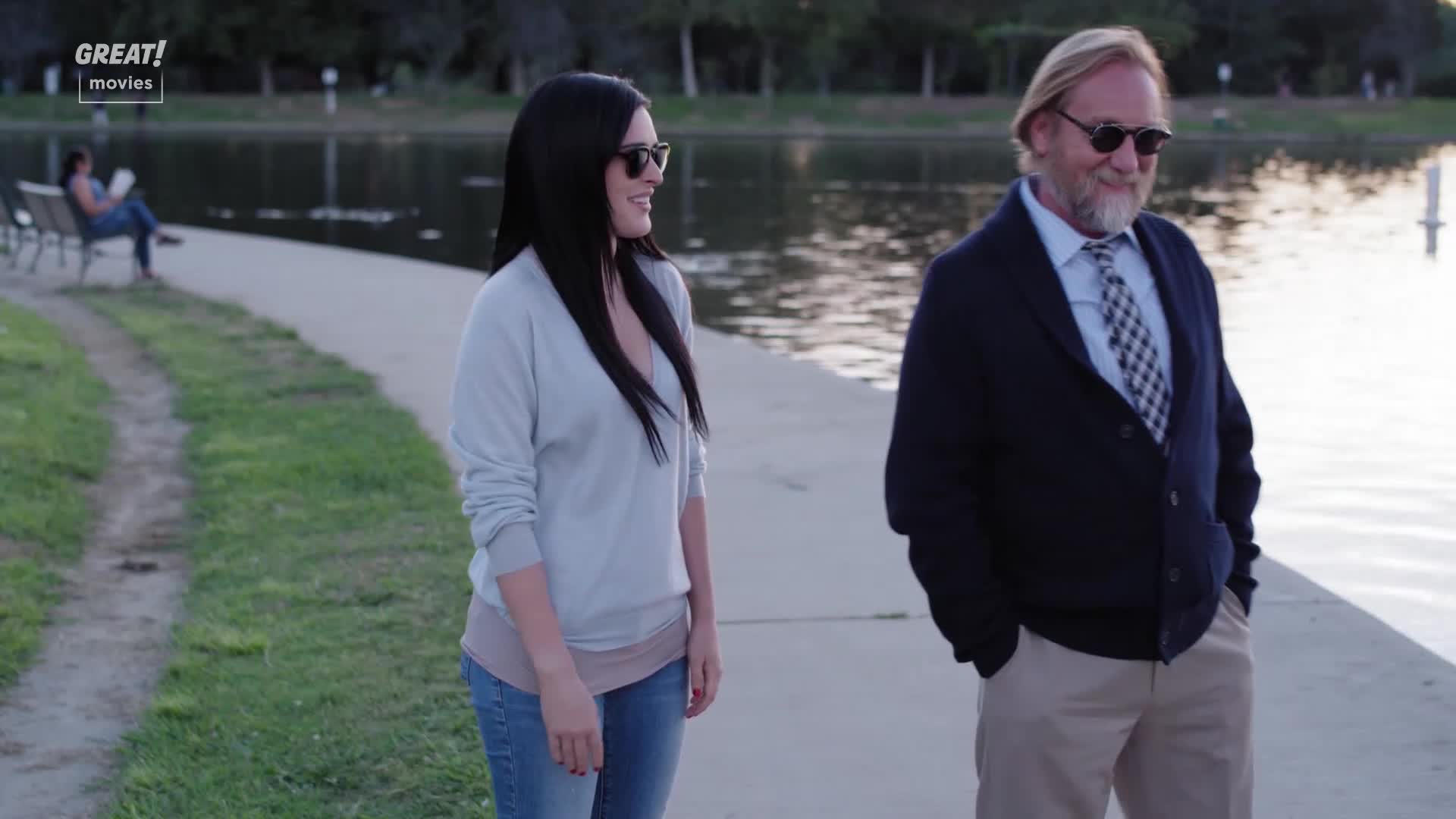A woman in a light sweater and jeans smiles as she walks alongside a man in a blazer and tie. They're near a lake, and a woman sits on a bench in the background, reading.
