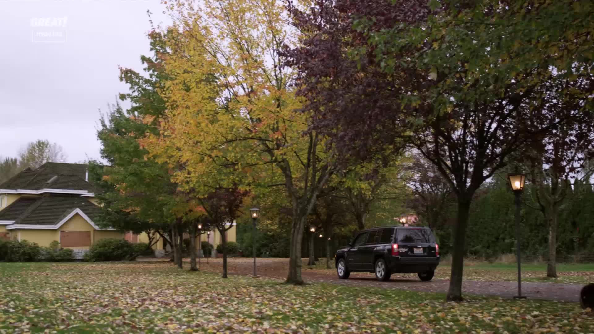 A dark SUV is slowly driving up the long, tree-lined driveway. The leaves on the trees are turning gold and red, and the overcast sky hints at a crisp autumn day.
