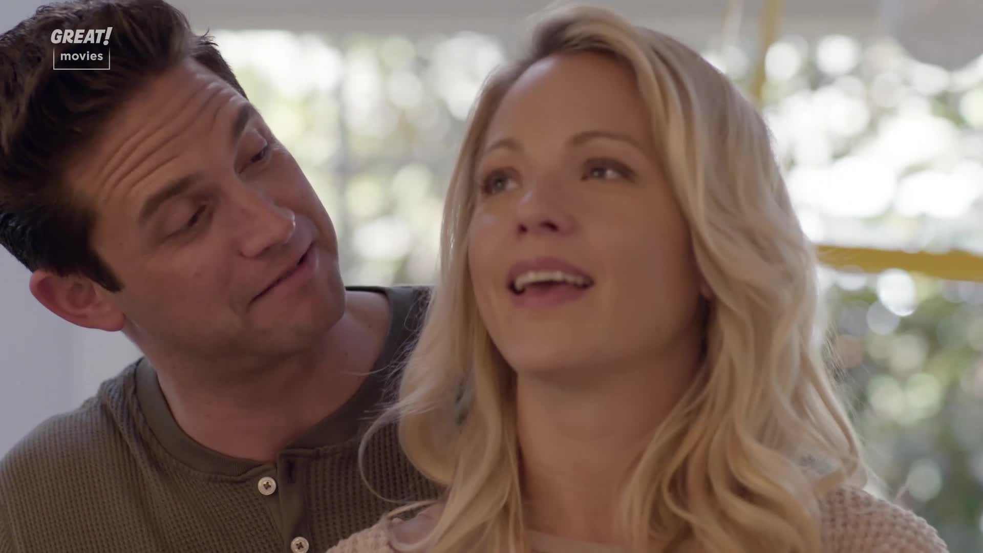 A woman with blonde hair is speaking, her mouth open as she looks up. A man is looking at her, smiling with his head tilted slightly.
