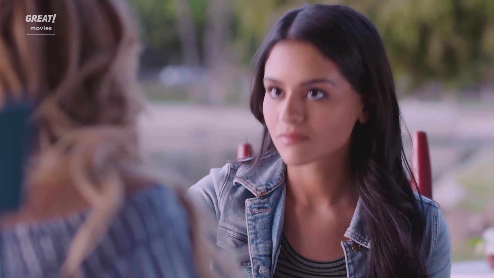 A young woman in a denim jacket is listening intently, her gaze fixed on someone out of frame. The light catches her dark hair and the serious expression on her face.
