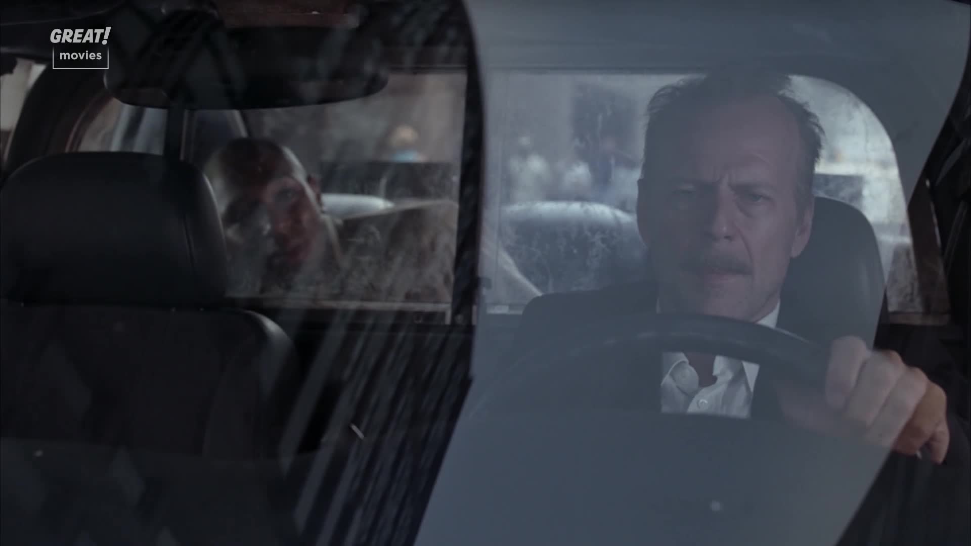 A man in a suit steers a car, his face focused, while in the back, another man stares out, his face bloodied. Rain streaks the windshield, obscuring the outside world.
