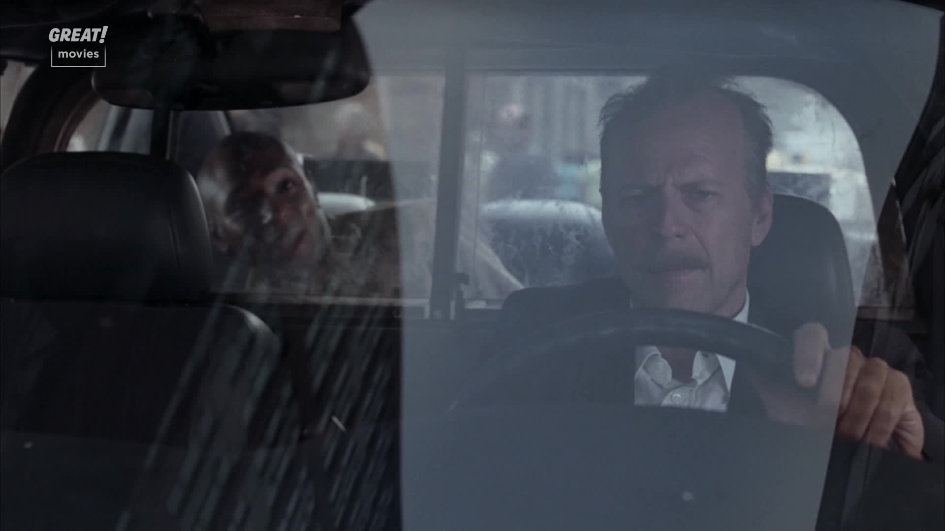 The man in the driver's seat grips the wheel, his face set in a grim expression. Through the rain-streaked window, another man peers in, his gaze intense.
