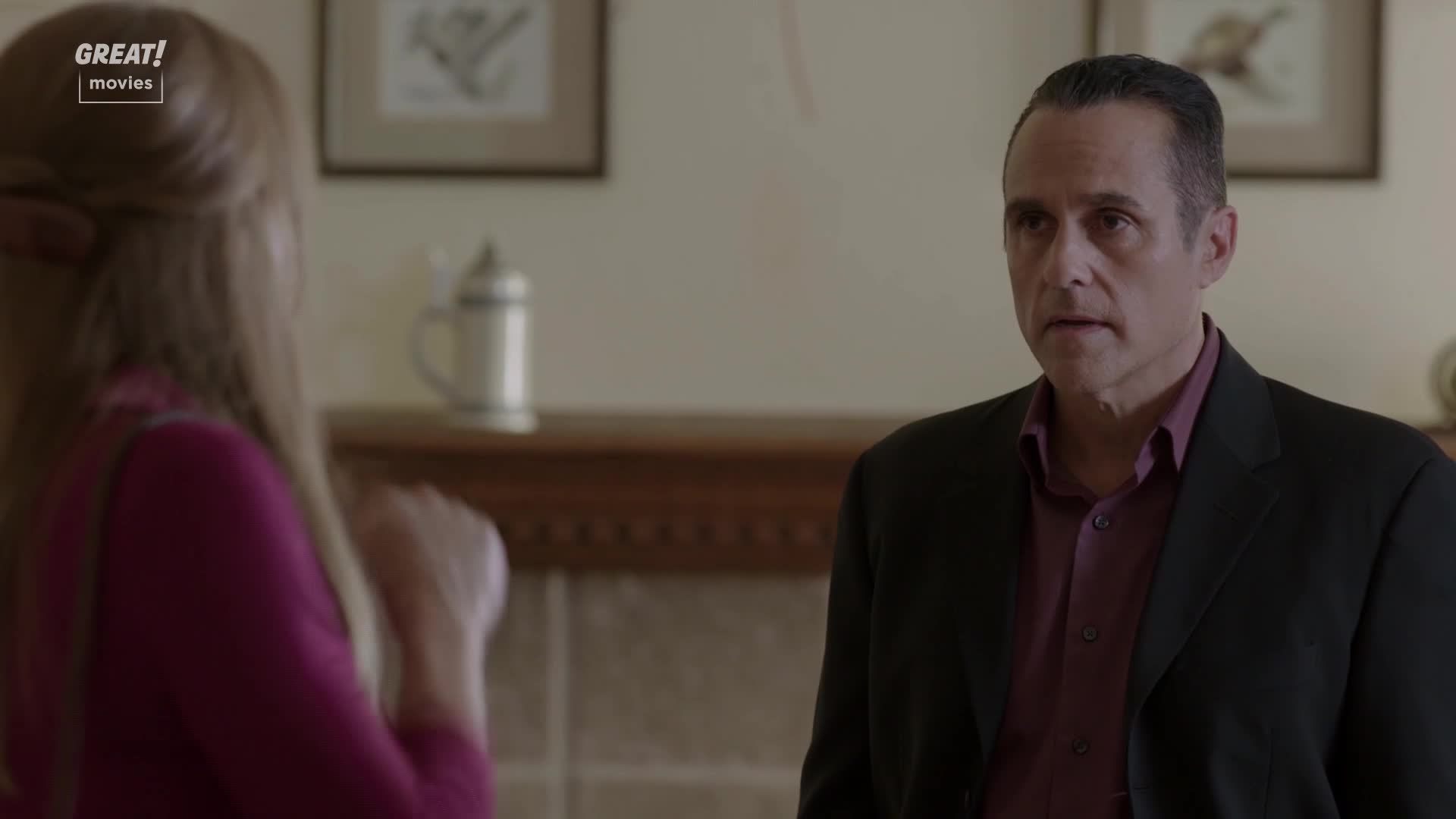A man in a dark suit stares intently at someone off-screen, his expression a mixture of concern and perhaps a bit of surprise. The woman in a pink top, partially visible, gestures with her hands, likely mid-conversation.

