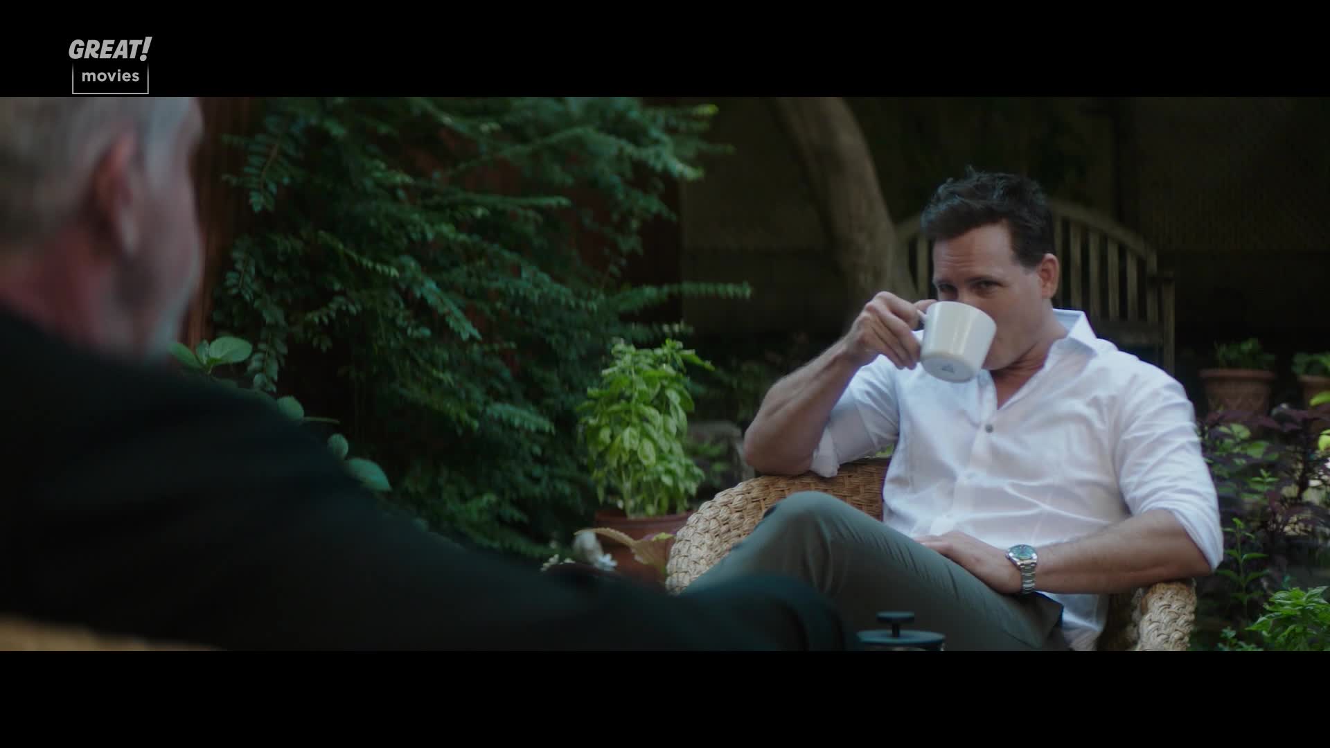 A man in a white shirt sips from a mug, seated in a wicker chair. Across from him, another man is partially visible, dressed in black.
