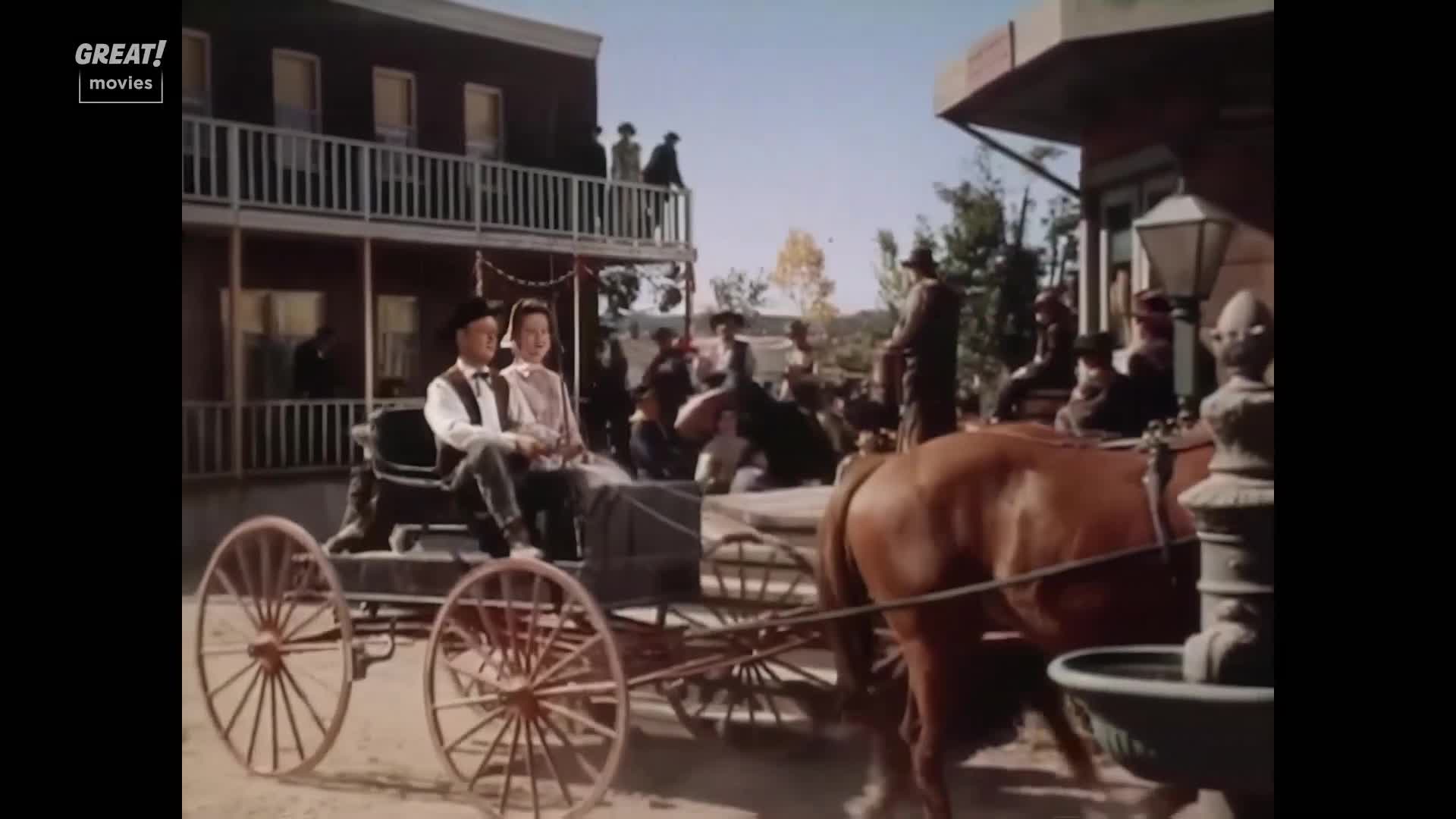 A horse-drawn carriage, carrying a couple, slowly rolls past the crowd.  People watch from the balcony of a wooden building, as if they're in a scene from a classic British film.

