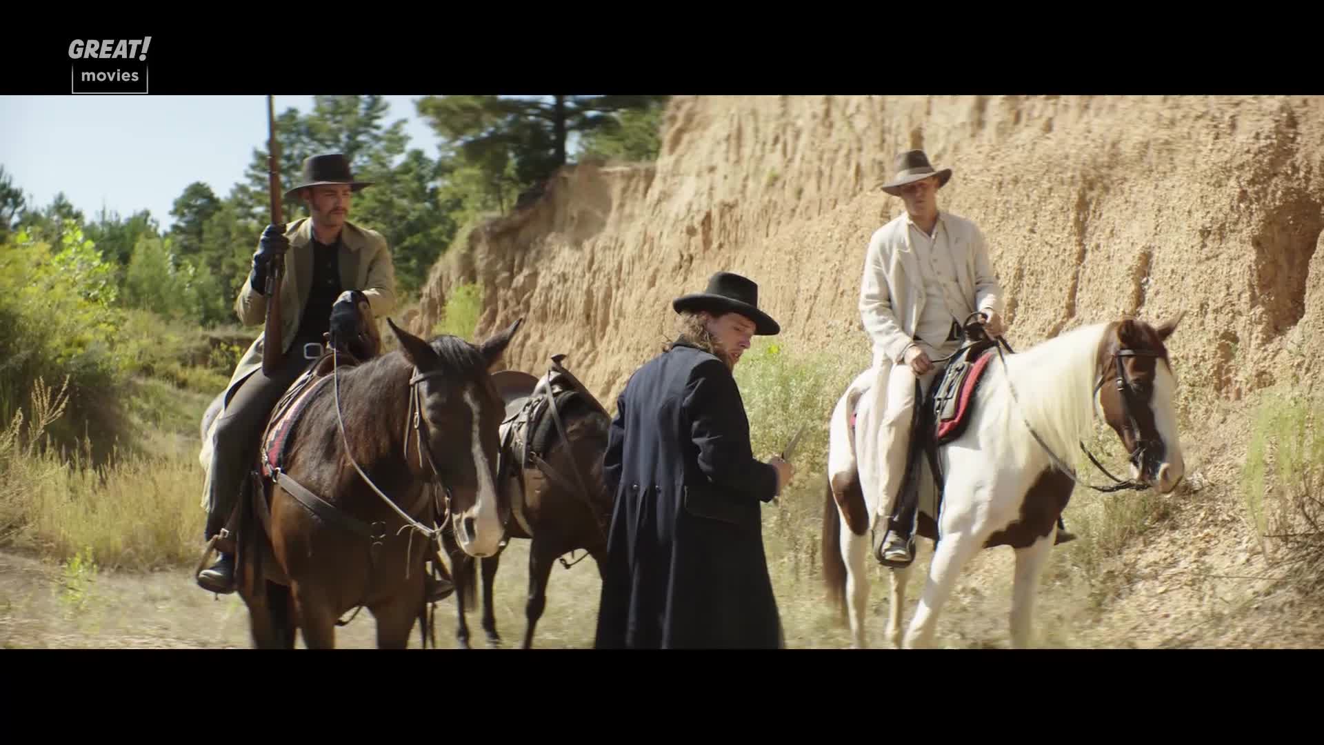 Three men on horseback are stopped on a dirt path, one holding a rifle. A fourth man in a long black coat stands beside them, looking at the riders.
