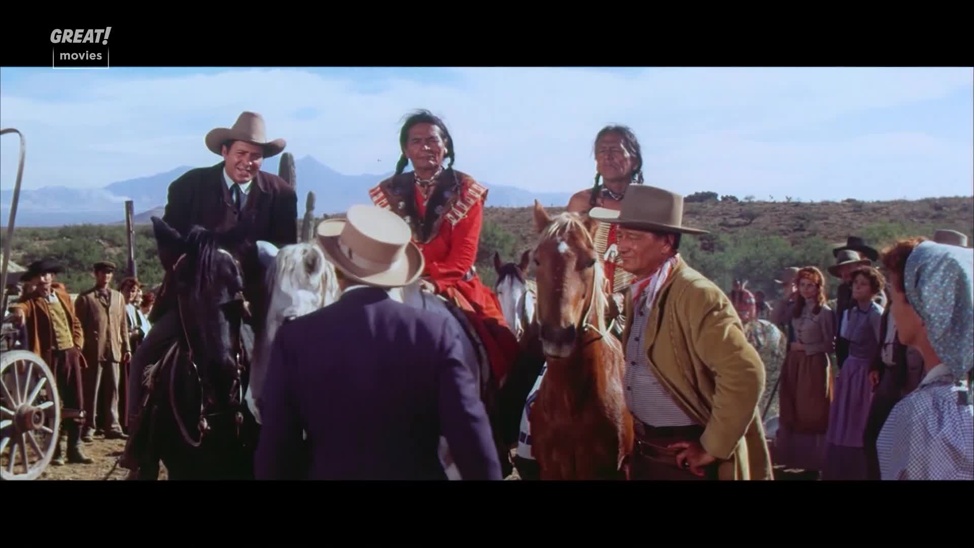 A group of people on horseback, including two Native Americans in traditional dress, are gathered in a dusty landscape. John Wayne, in a tan jacket, stands next to a brown horse, talking with a man in a suit.
