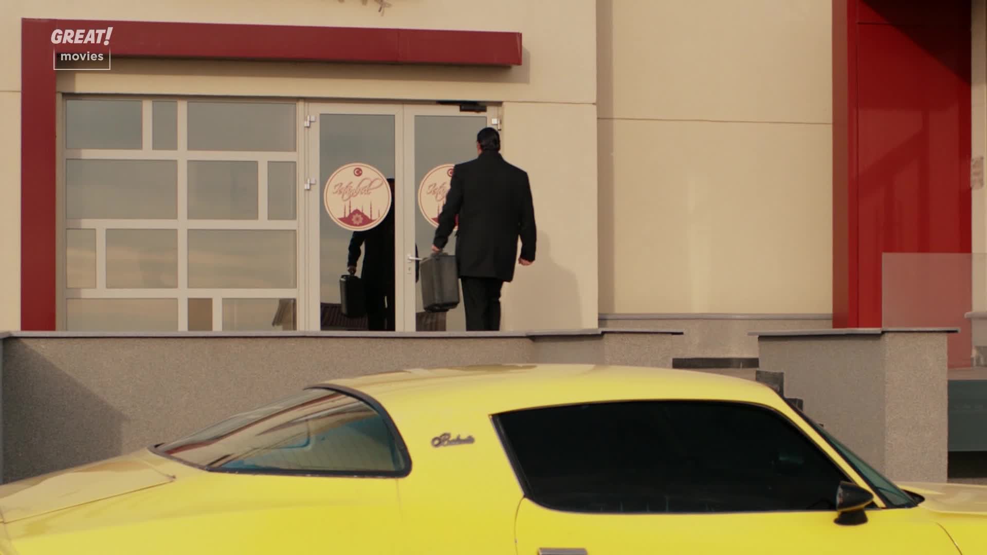 A man in a dark suit exits the building, a briefcase in his hand. The bright yellow car, parked in front, is a striking contrast to the man's somber attire.
