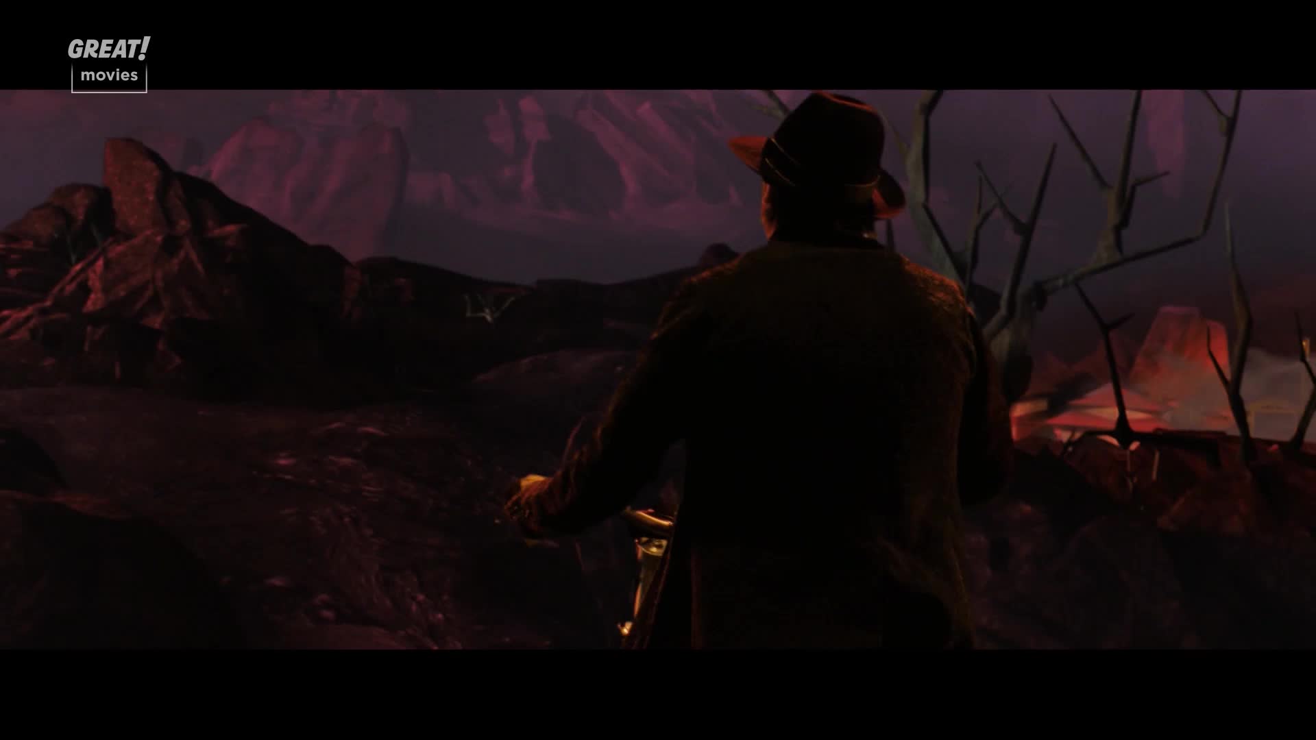 A man in a fedora slowly cycles forward, the bicycle's handlebars barely visible. The landscape is a desolate, reddish terrain, with jagged rock formations and skeletal trees. 
