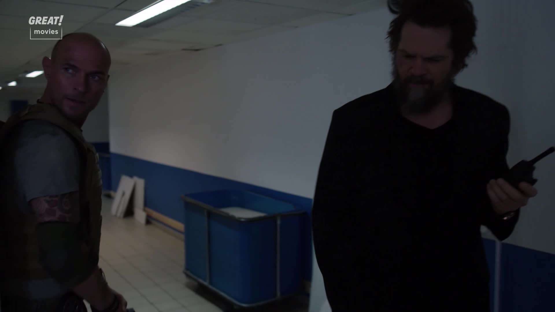 The man in tactical gear glances back over his shoulder, while the other man, holding a walkie-talkie, continues down the hallway. They're in a drab, institutional space, the kind you see in a lot of United Kingdom thrillers.
