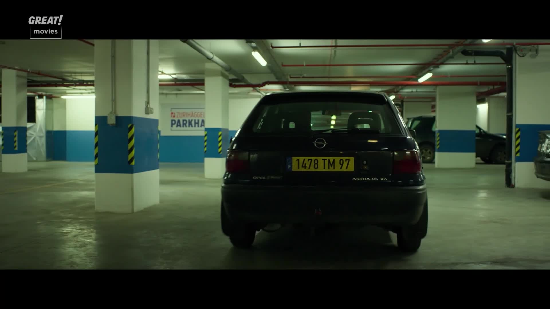 A black Opel Astra sits in a well-lit parking garage, its yellow license plate reflecting the overhead lights. The car is parked, but it could be ready to make a quick getaway.
