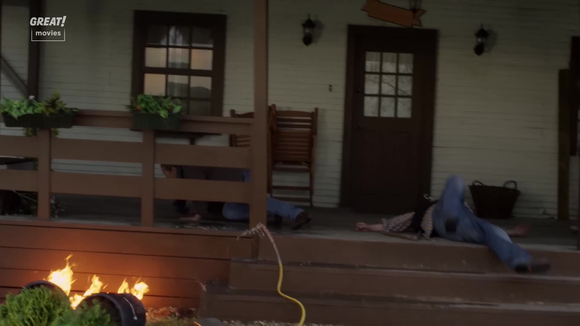 A man lies sprawled on the porch steps, legs splayed out. Flames lick at a barrel near the edge of the porch, and another person is on the porch.
