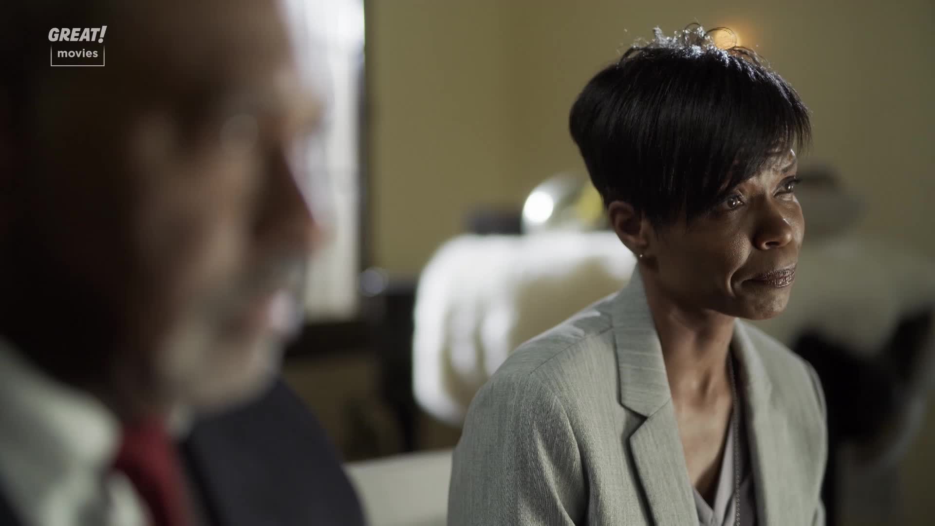 A woman in a grey blazer listens intently, her gaze fixed off-screen. Across from her, a blurred figure in a suit and red tie is talking, likely in a scene from a GREAT! Movies film.
