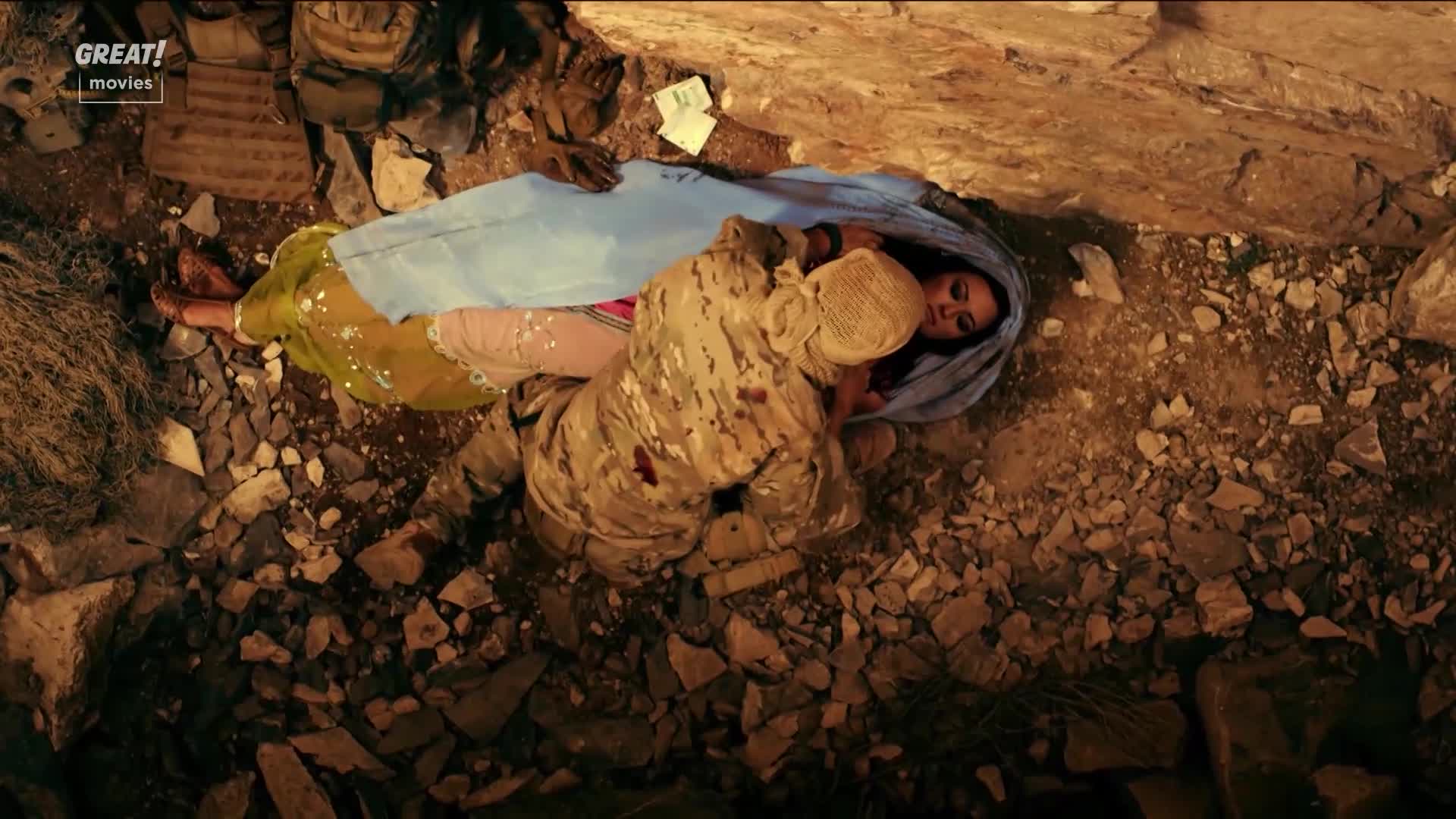 A woman in a blue headscarf lies on the ground, her colorful clothing visible. A soldier in camouflage kneels over her, his uniform stained with blood.
