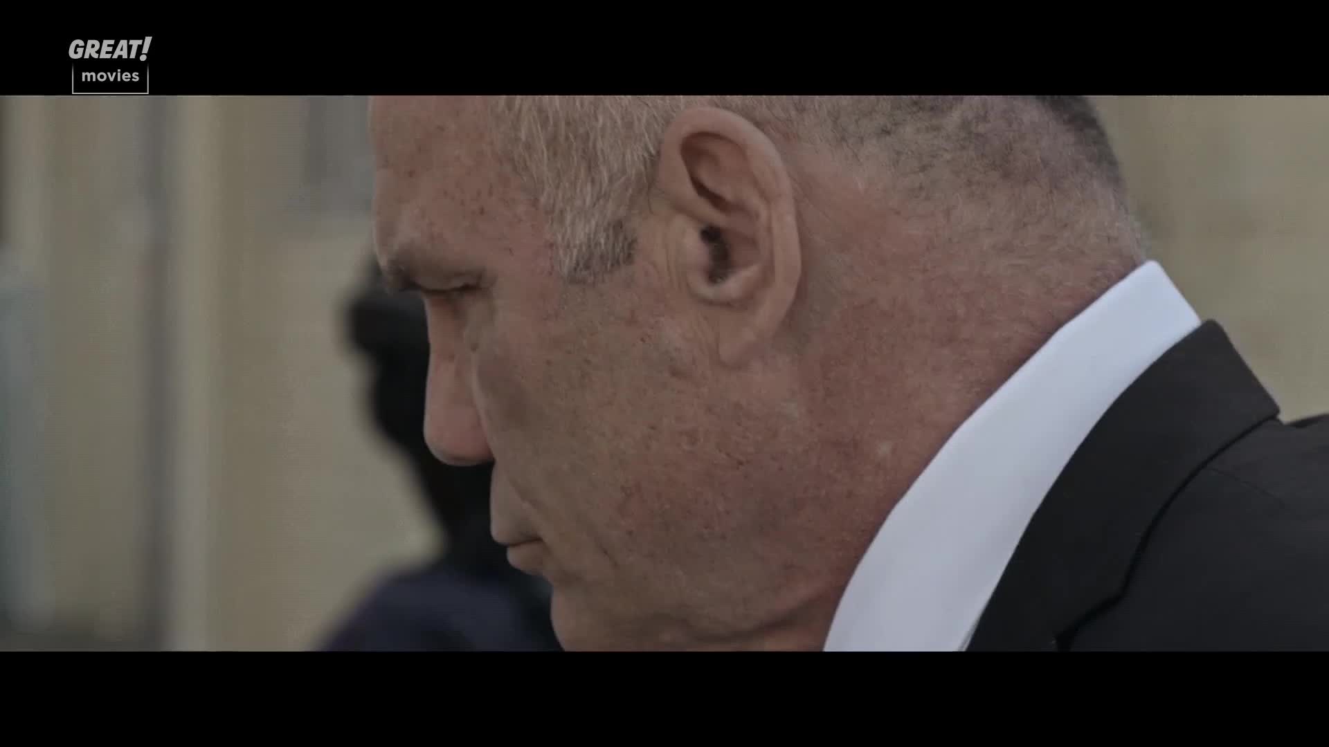 A man in a dark suit is looking down, his face in shadow. The GREAT! movies logo is in the upper left corner, and the scene appears to be from a film set in the United Kingdom.
