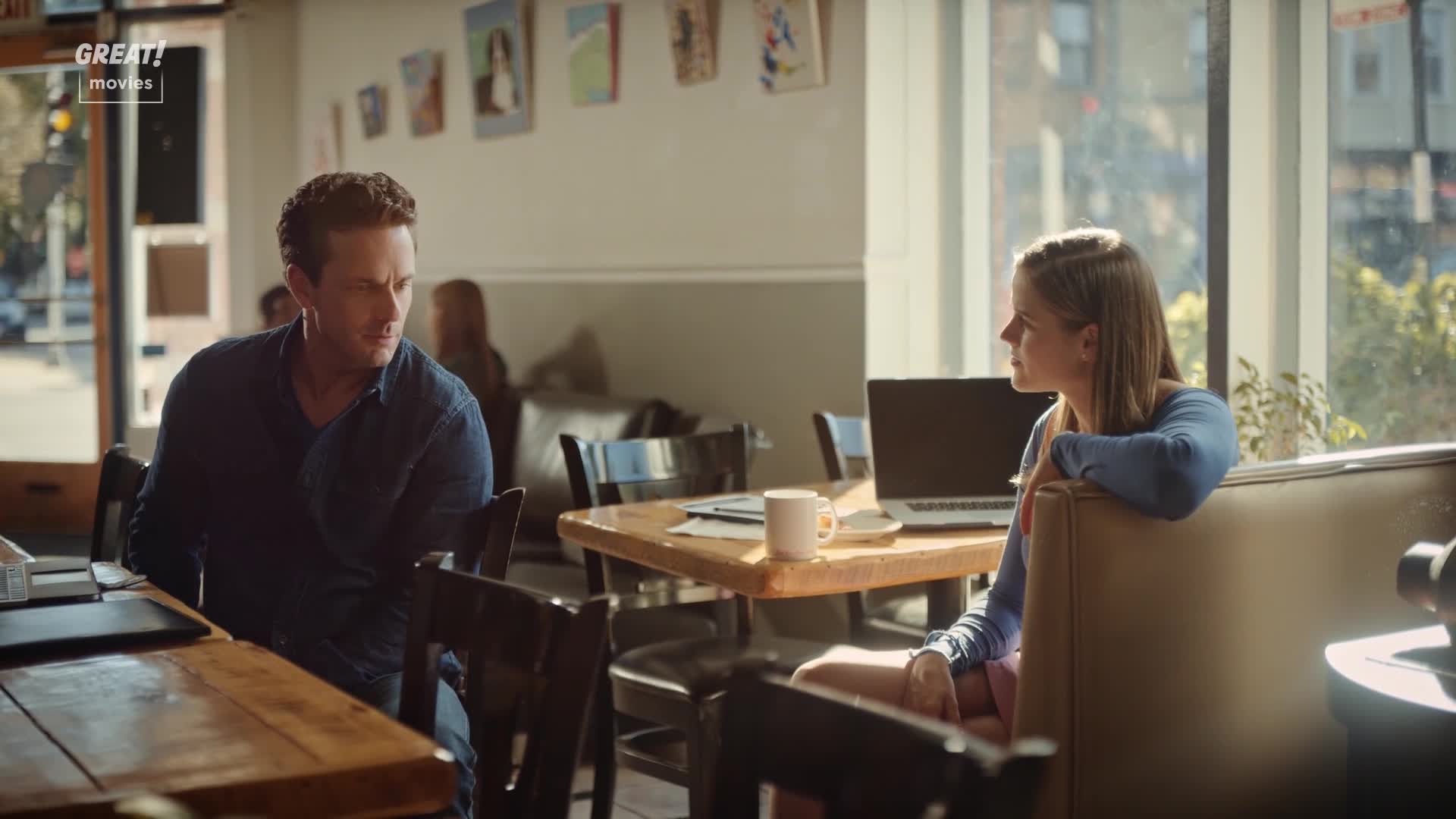 A man sits at a table, looking thoughtful, while a woman leans back, gazing out the window. Sunlight streams in, illuminating the cafe, a scene that wouldn't be out of place in a GREAT! movie.
