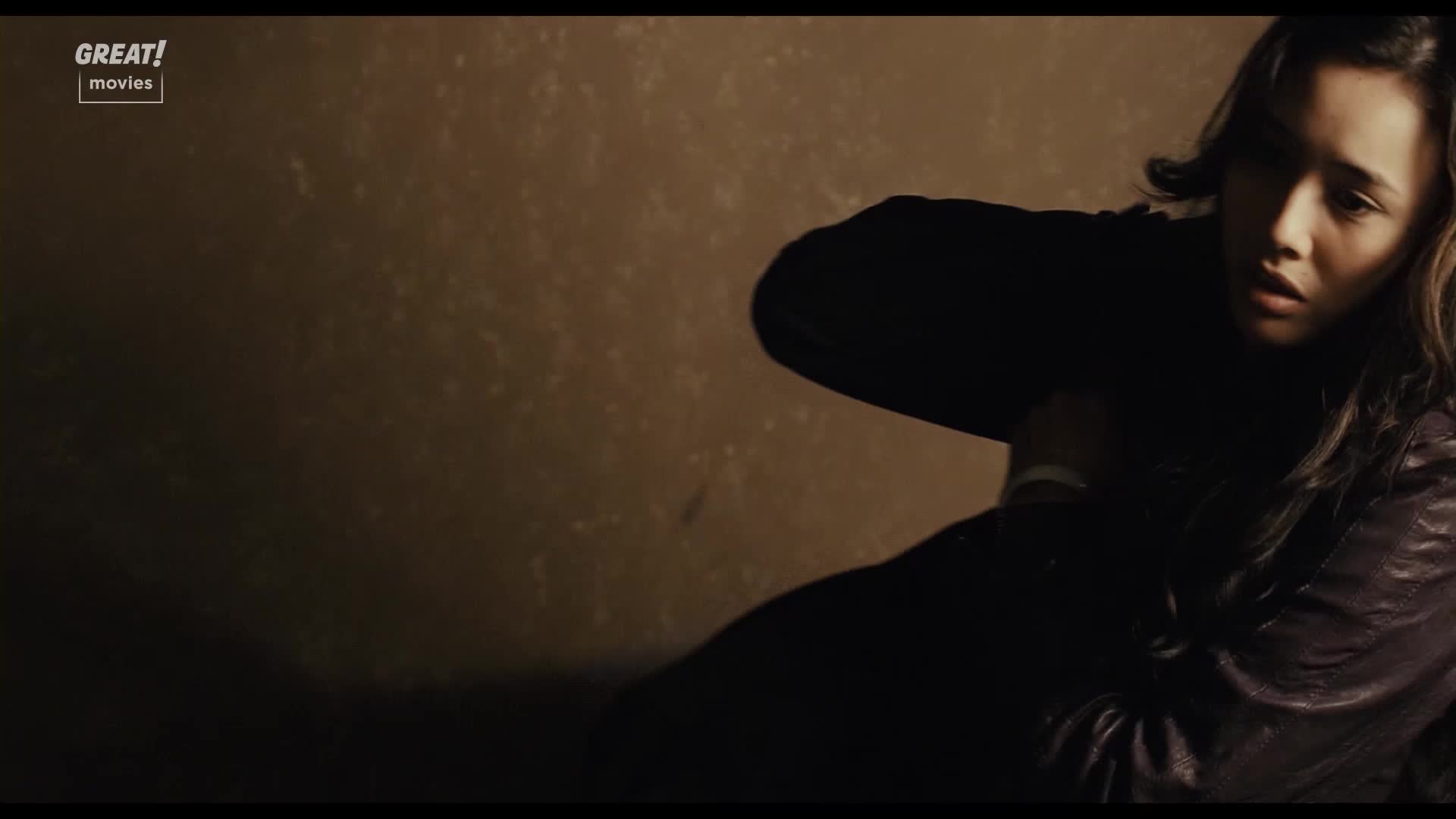 A woman is crouched, her face etched with worry. She's wearing a dark jacket, her arm bent as if protecting herself.
