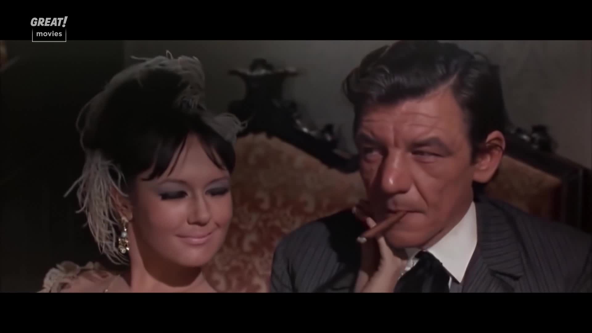 A woman in a feathered headdress smiles, her hand gently holding a cigar for the man beside her. He puffs on it, eyes narrowed, in a scene that feels straight out of a classic British film.
