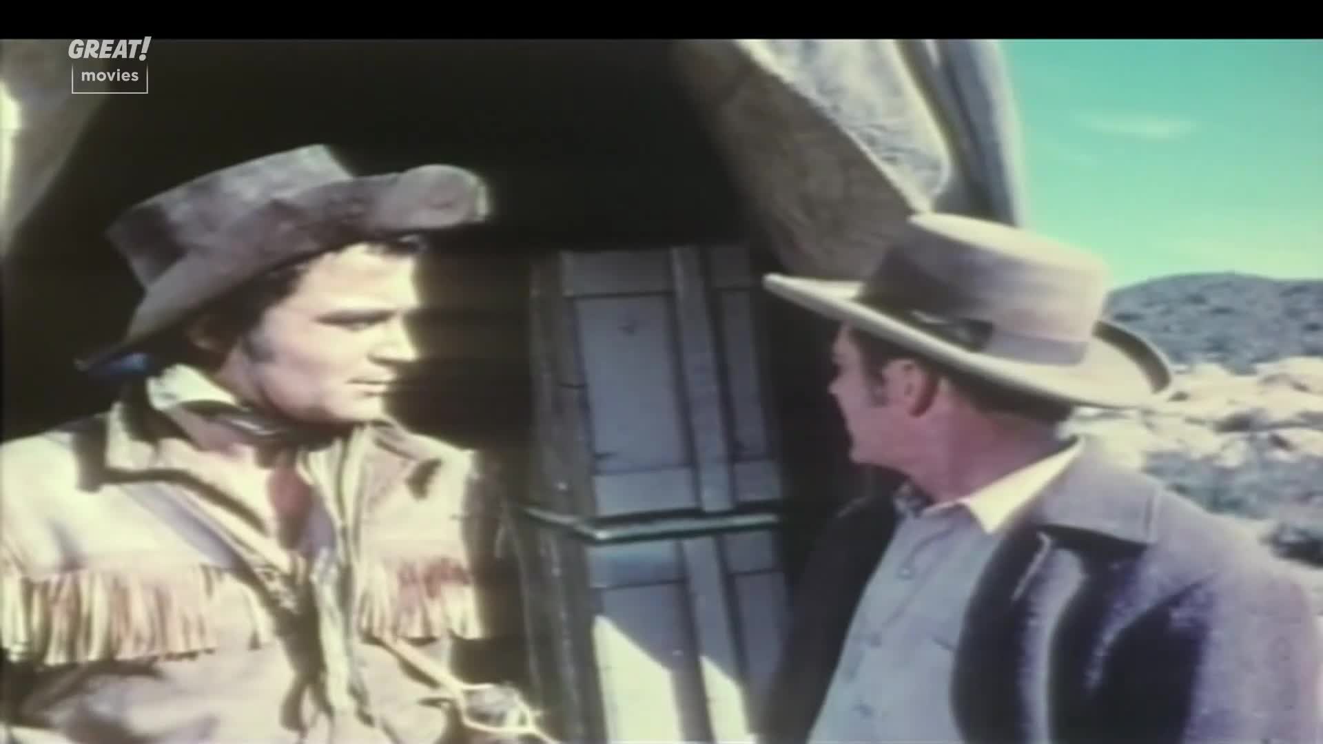 Two men, both in hats, are talking near a wagon. The man on the left, in a fringed jacket, seems to be listening intently to the other, who is turned slightly away.
