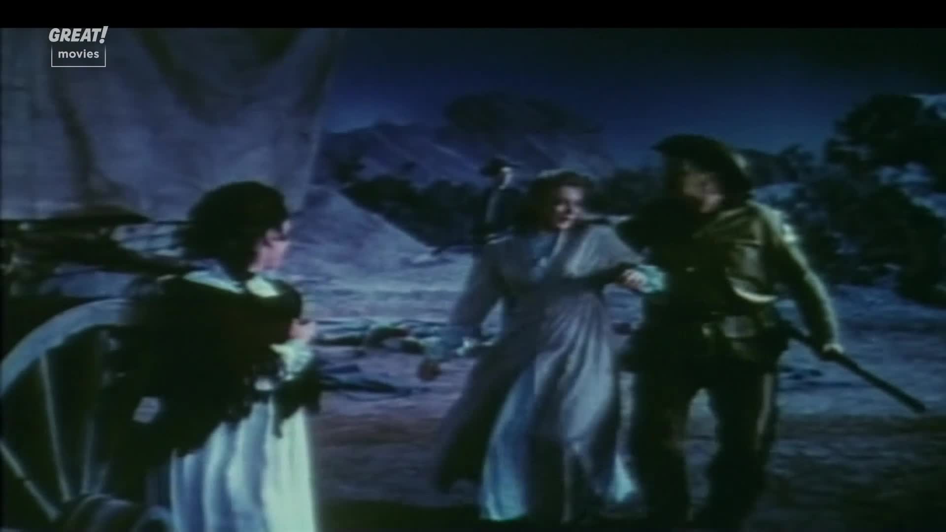 A woman in a long, light-colored dress is being led by a man in a military uniform, who holds a weapon. A third woman, dressed in dark clothing, watches them from near a wagon wheel.
