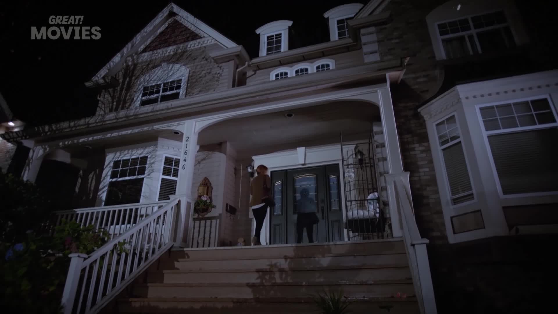 A couple stands on the porch of a large house, illuminated by the porch light. The front door is ajar, and the house number, 21646, is visible on the side of the house.
