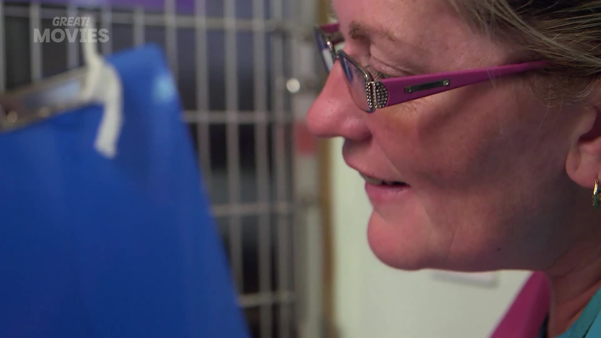A woman with glasses and a pink shirt is looking down, her gaze focused on something just out of view. Beside her, a bright blue object is partially visible, held in place by a metal frame.
