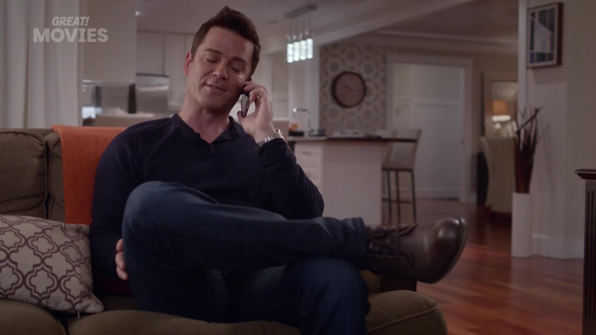 A man in a dark sweater lounges on a sofa, phone pressed to his ear. He's smiling, his legs crossed, boots resting on the cushions.
