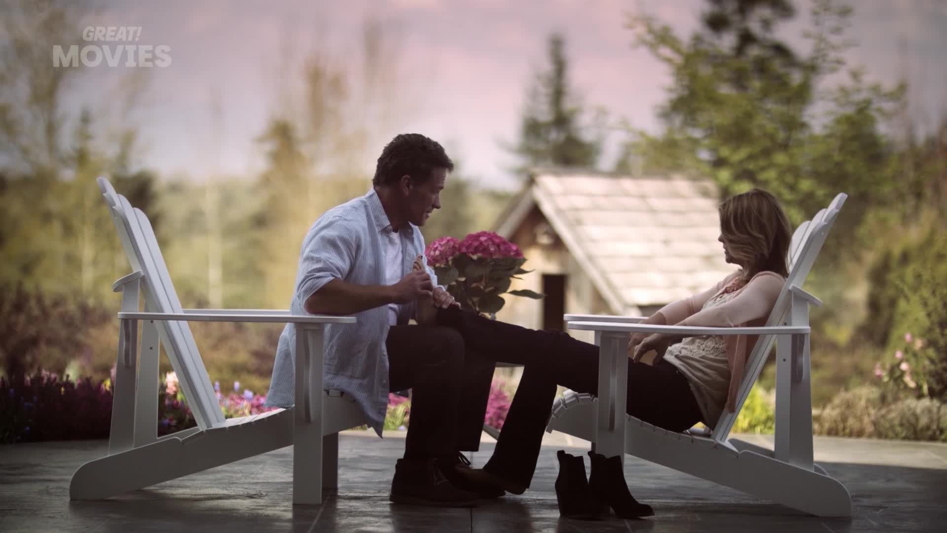 A man kneels, gently massaging a woman's foot as she sits in a white chair. They're outside, with a cottage and a flower arrangement nearby, a scene that feels like a romantic moment from a GREAT! Movies film.

