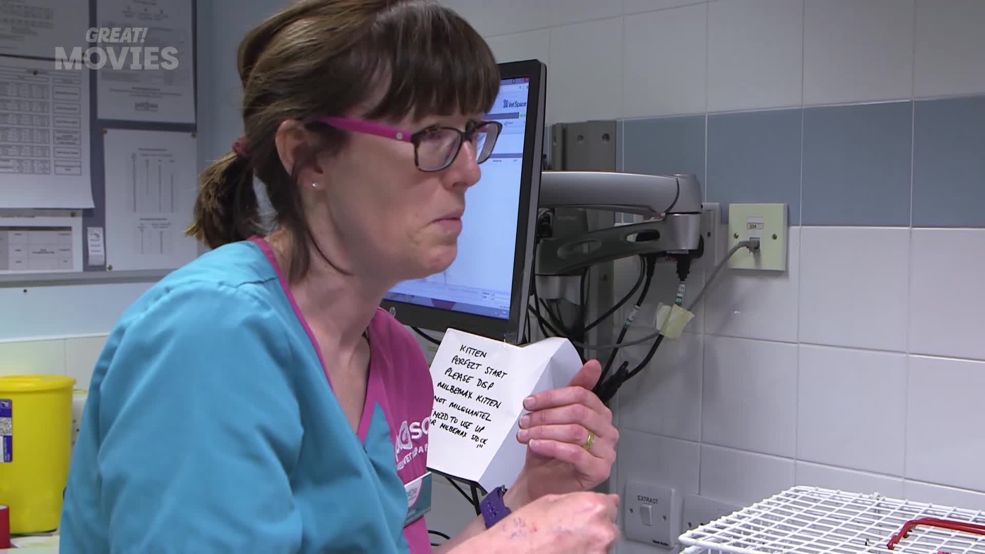 A woman in scrubs, wearing glasses, is holding a folded piece of paper, reading the notes scrawled on it. She's in a veterinary clinic, likely from a GREAT! MOVIES film.
