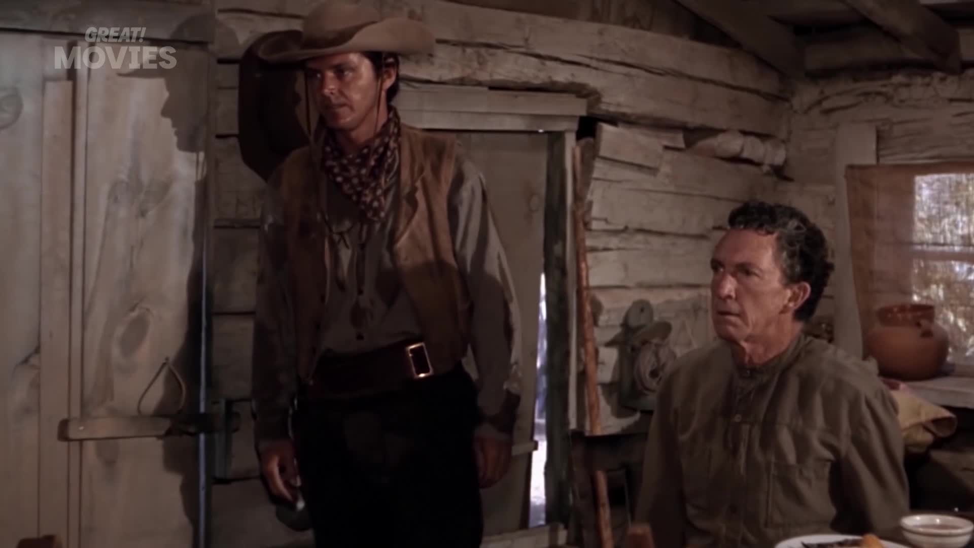 A cowboy in a hat stands in the doorway, looking intently at the man seated at the table. The older man stares back, his face etched with worry.
