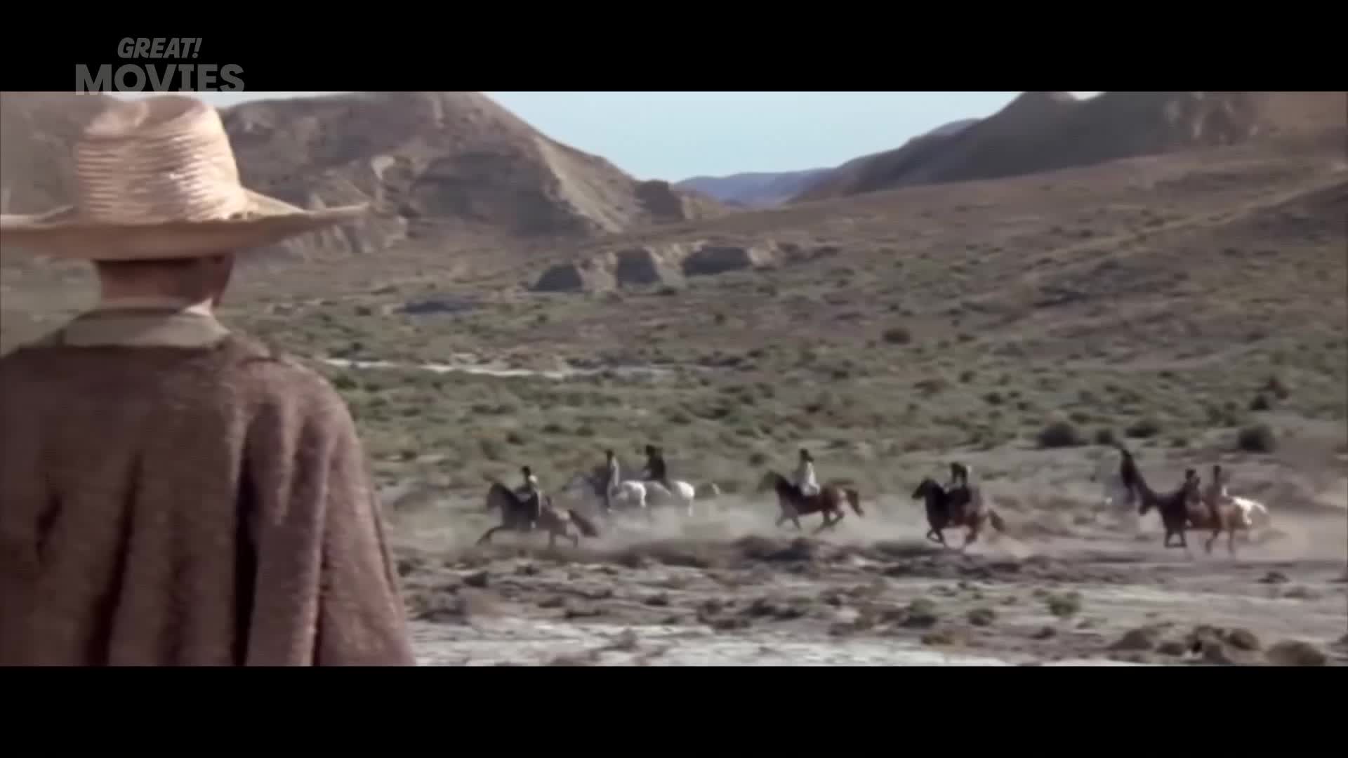 Dust billows as a group of riders on horseback gallop across the arid landscape. A man in a wide-brimmed hat watches them from the foreground.
