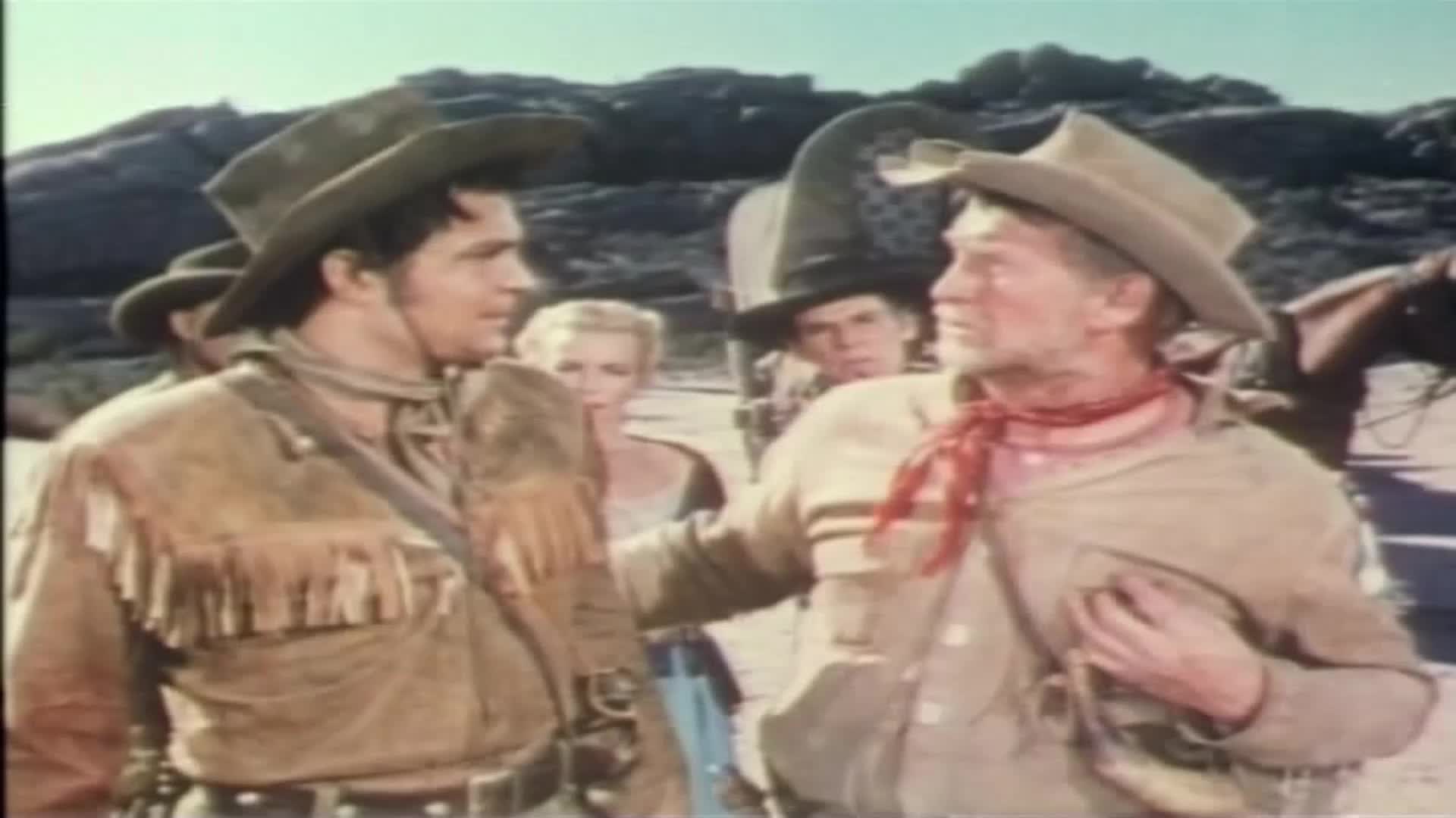 The older man, wearing a red neckerchief, puts a hand on the younger man's shoulder. A woman with blonde hair stands behind them, watching the exchange.
