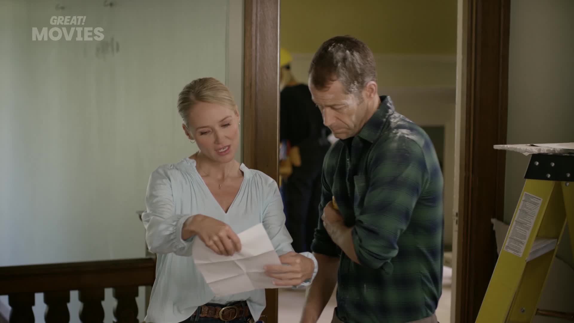 A woman in a light blue blouse gestures at a folded paper, explaining something to the man beside her. He's wearing a green plaid shirt and looks intently at the document.
