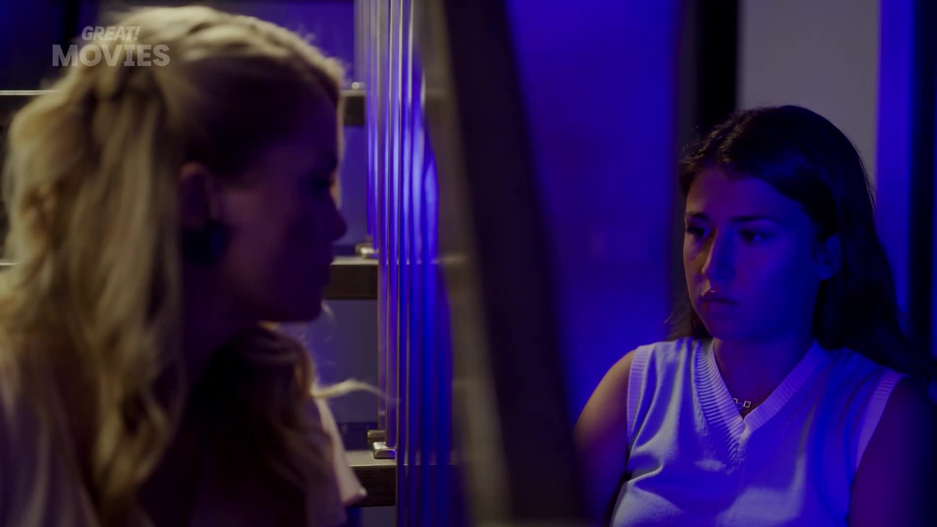The woman with blonde hair is turned away, speaking to the other woman who sits facing forward, looking concerned. The scene is lit with a deep blue hue, suggesting a late-night conversation.
