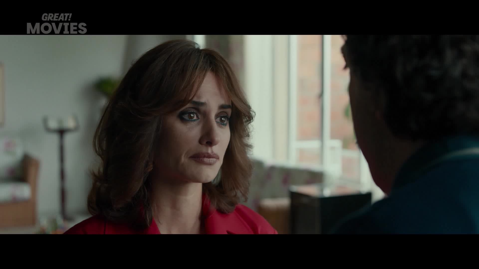 Her eyes are fixed on the other person, her face etched with a mix of sadness and concern. The red of her jacket pops against the muted background of a room, perhaps a scene from a UK-made film.
