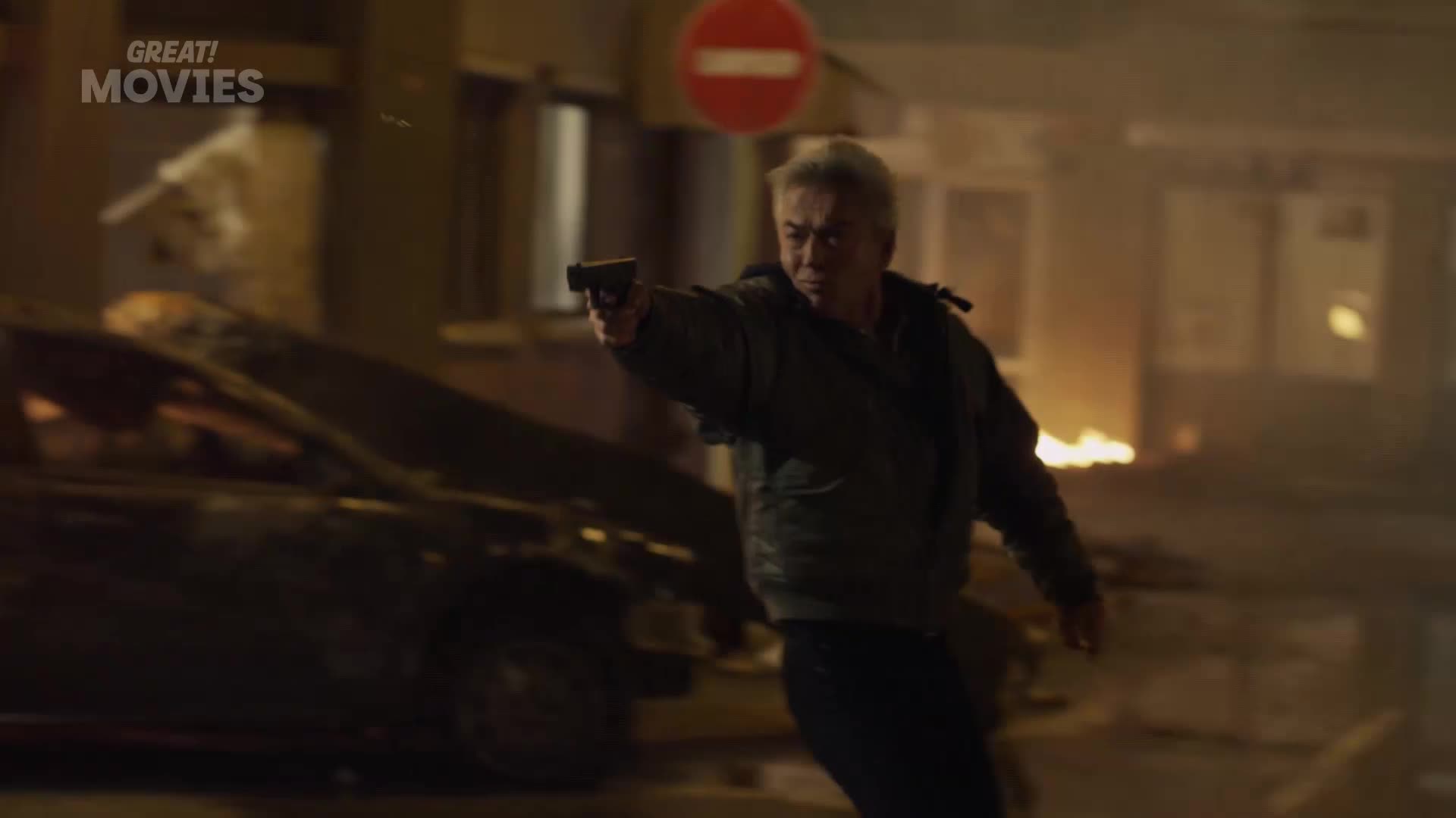 A man with silver hair holds a gun, aiming it ahead. Behind him, a car looks wrecked, and a fire burns in the distance.
