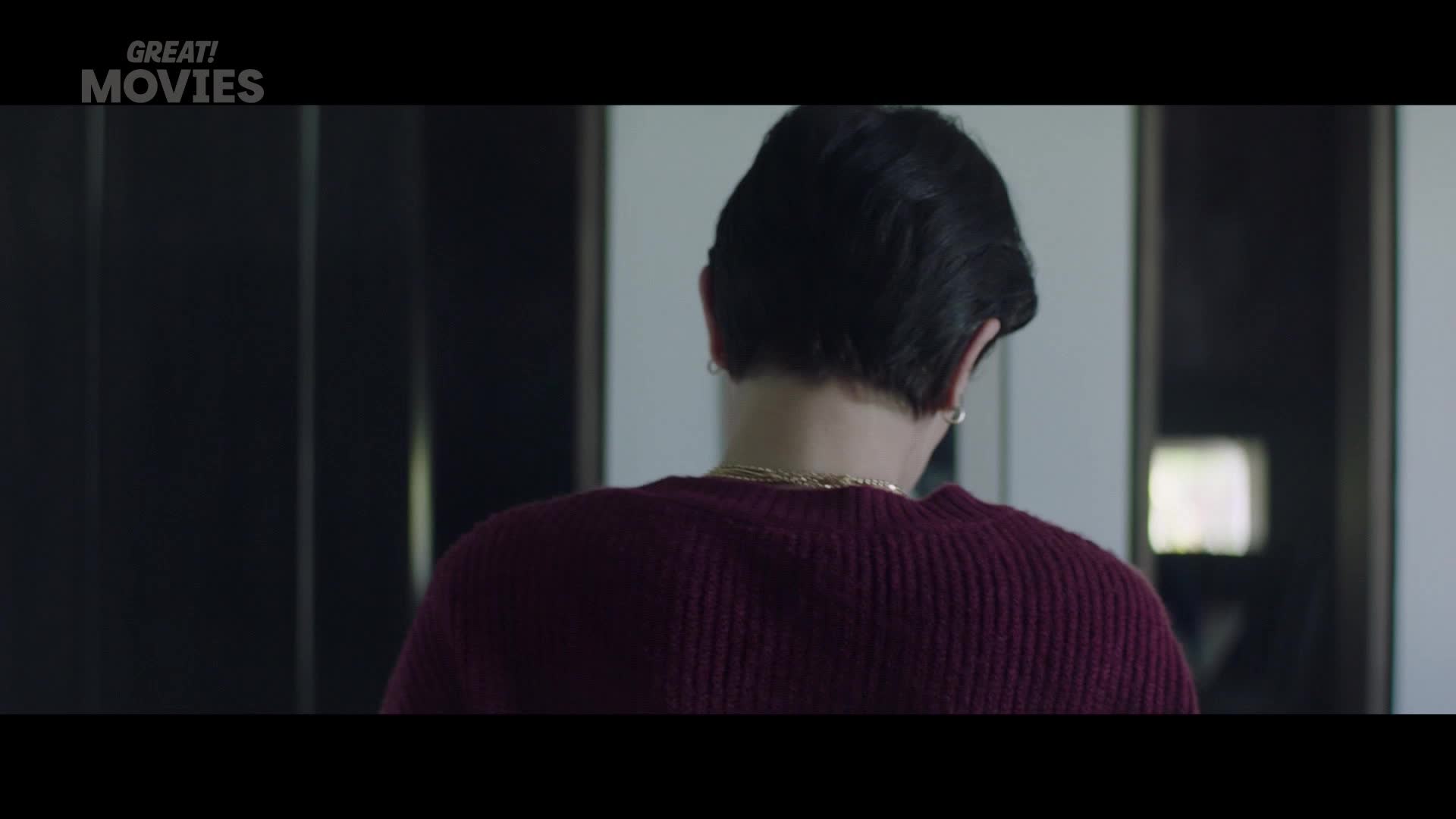 A person with dark hair and a gold chain turns away, their burgundy sweater a rich texture against the muted background. This scene, like many on GREAT! Movies, feels like a quiet moment before something unfolds.