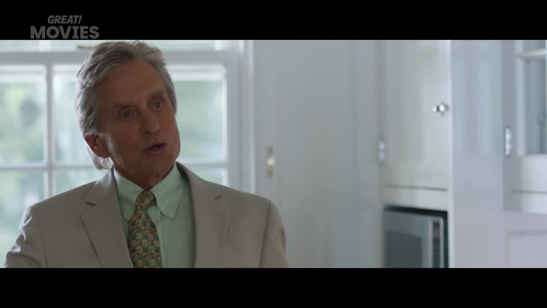 A man in a suit is talking. He's in a kitchen with white cabinets.