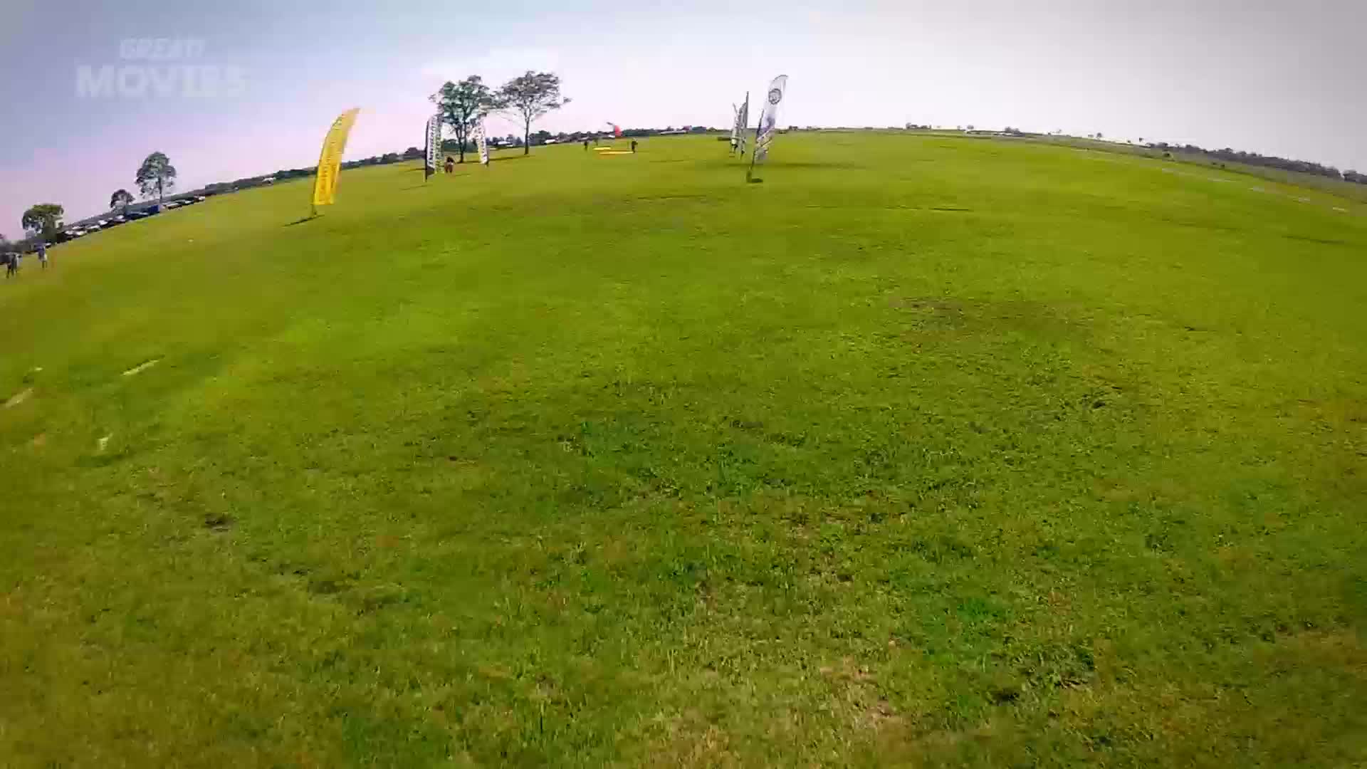 A bright yellow flag stands tall on the grassy slope, marking a point on the course. People are scattered across the field, some walking, others appearing to be setting up for an event.