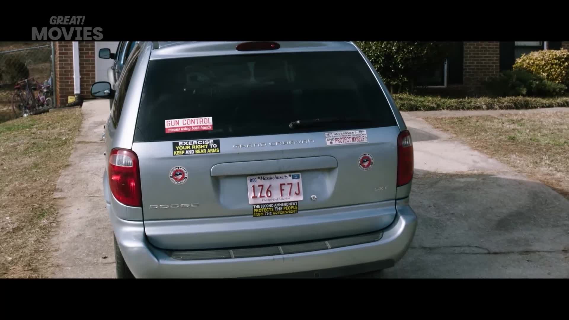 A silver Dodge Caravan is parked on a driveway. Its rear bumper is adorned with several stickers, including one that reads "GUN CONTROL" and another stating "EXERCISE YOUR RIGHT TO KEEP AND BEAR ARMS."