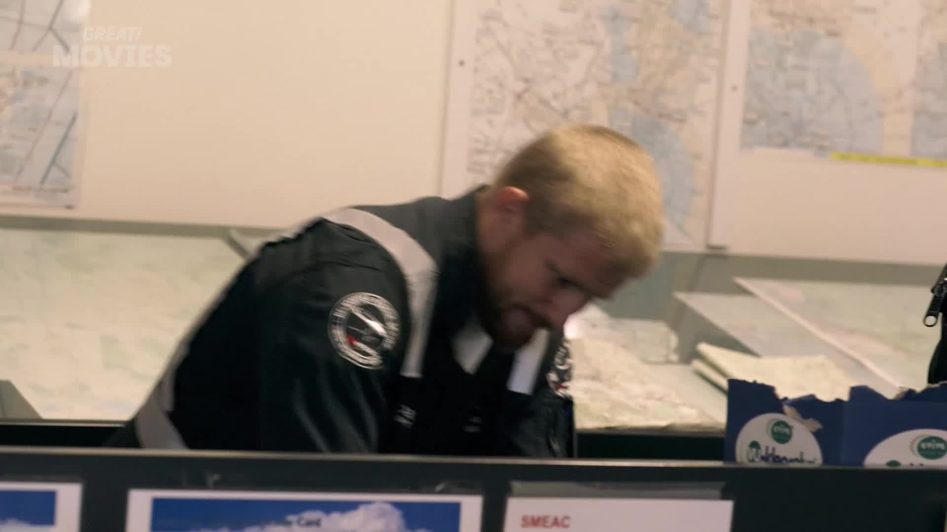 A man in a dark uniform with grey stripes leans over a desk, his blond hair falling forward. Maps are spread out on the wall behind him, and a blue box with white lettering sits on the desk.