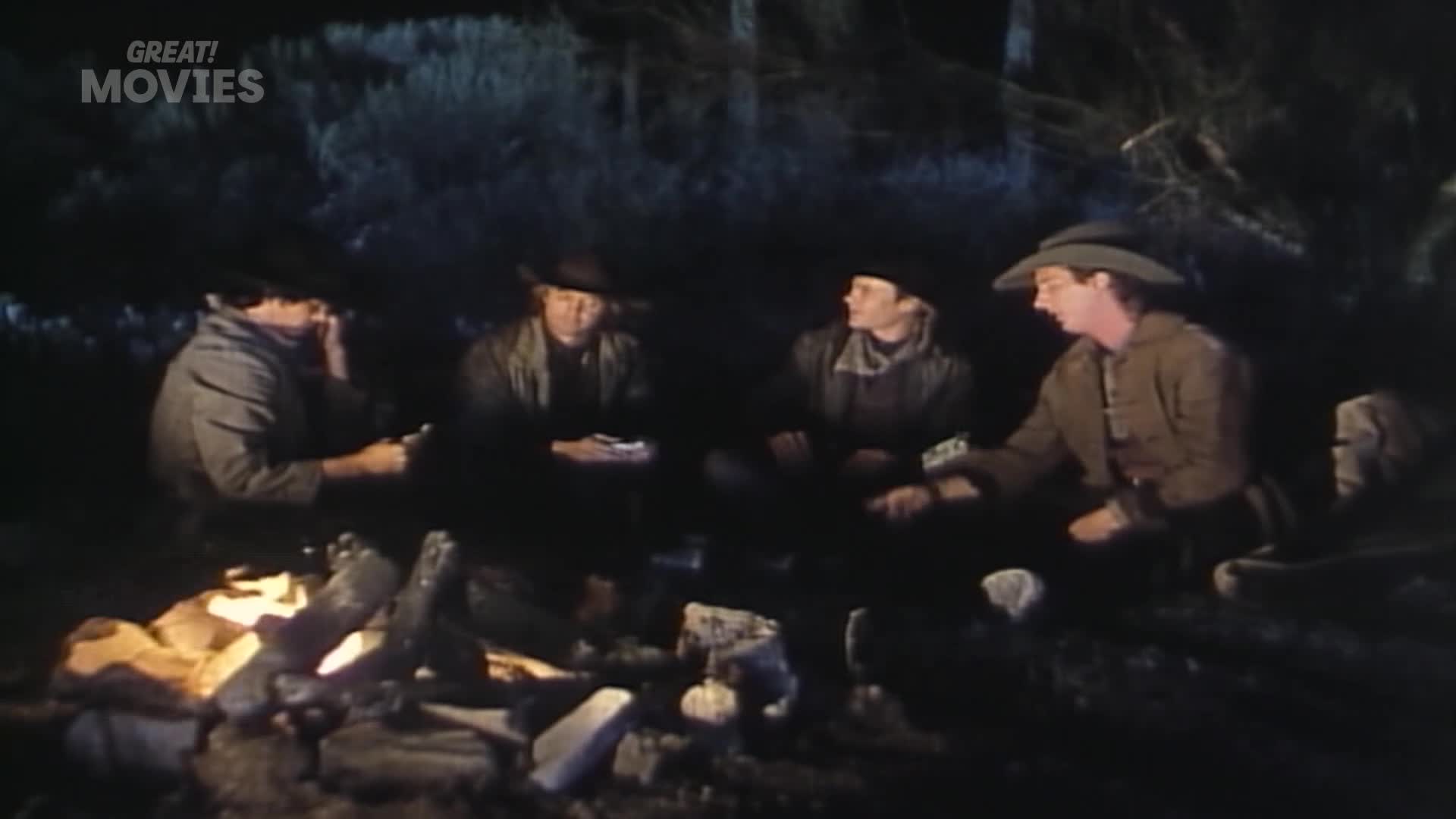 Four men huddle around a campfire, their faces illuminated by the flickering flames. They appear to be playing cards in the dim light of the woods.