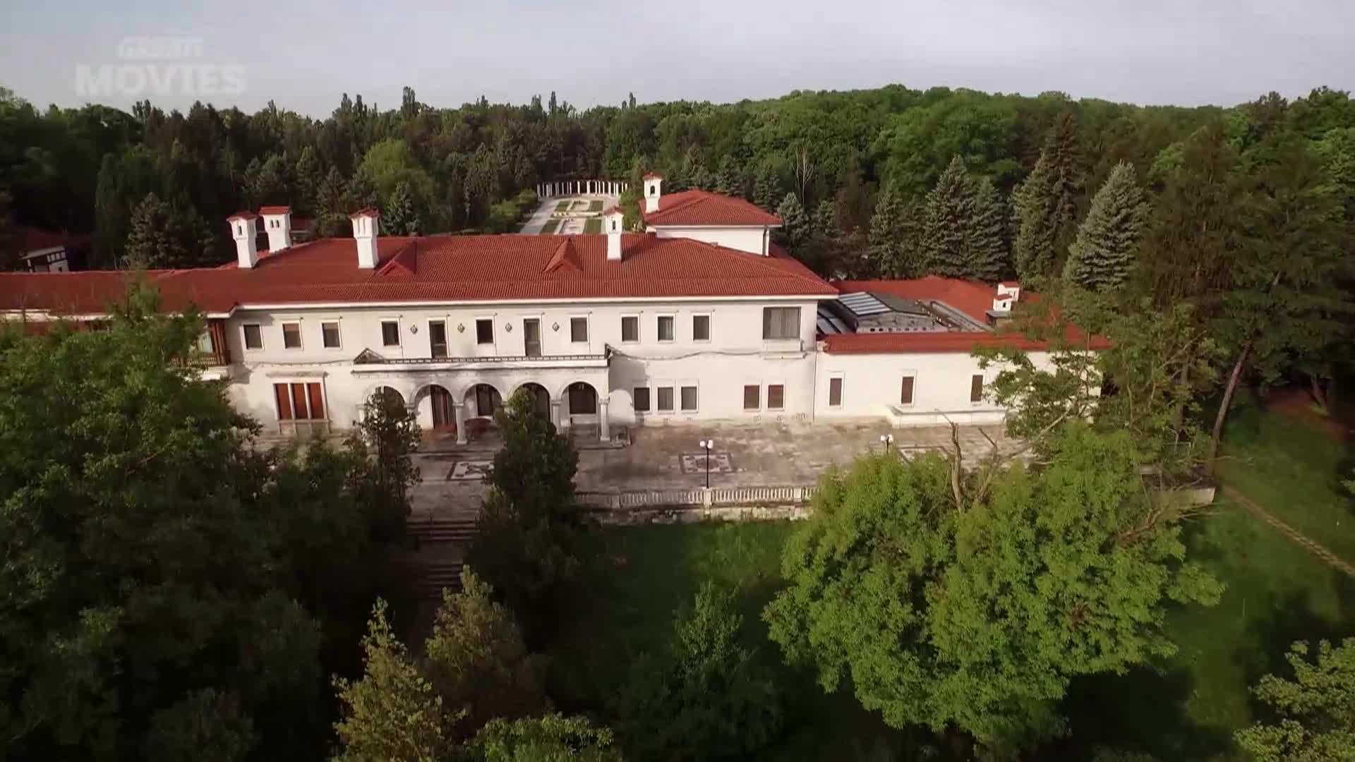 A grand white manor with a red-tiled roof sits nestled amongst lush green trees. The manor's architecture, with its arched walkways and numerous windows, suggests a stately home that could feature in a GREAT! movie.