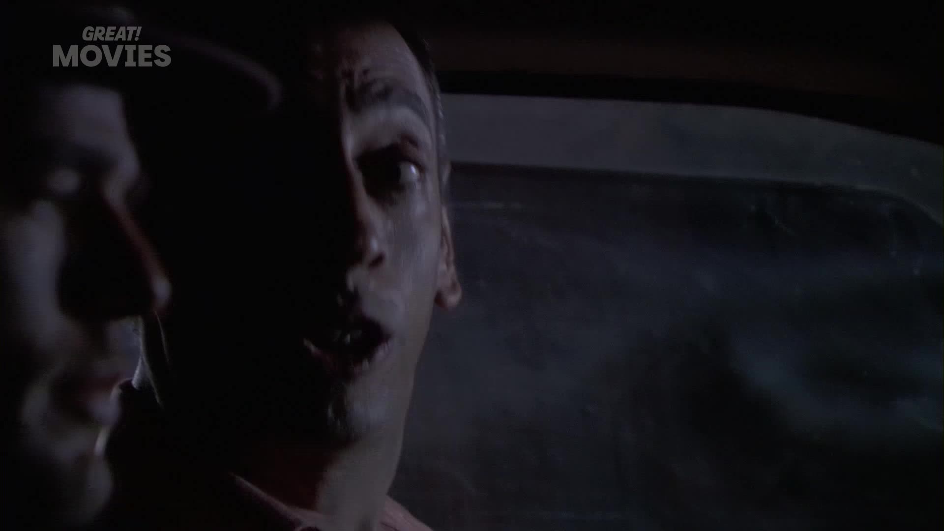 A man's face, illuminated by a faint light, is pressed close to the window of a car. He looks out with wide, startled eyes.
