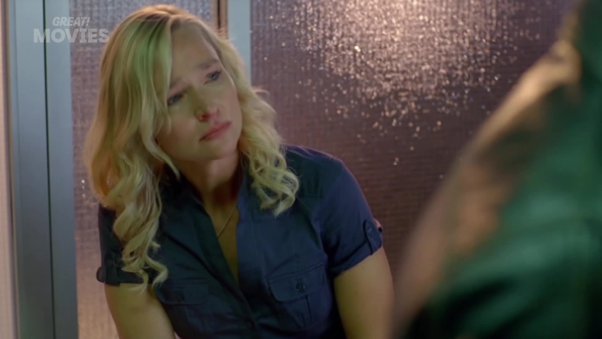 A woman with blonde, wavy hair looks down with a troubled expression. Rain streaks down a textured glass panel behind her.
