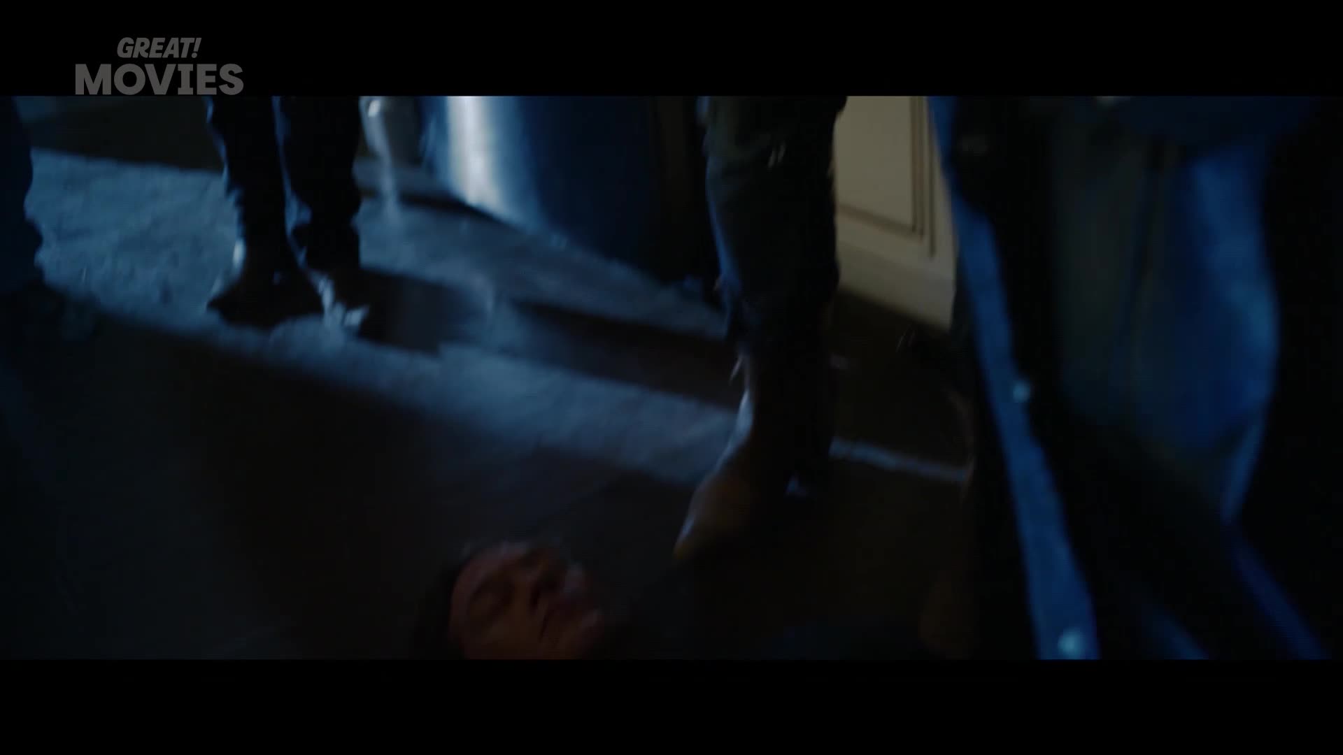 A man lies on the floor, his face pale in the dim light. Several figures in dark clothing stand over him, their boots casting long shadows.