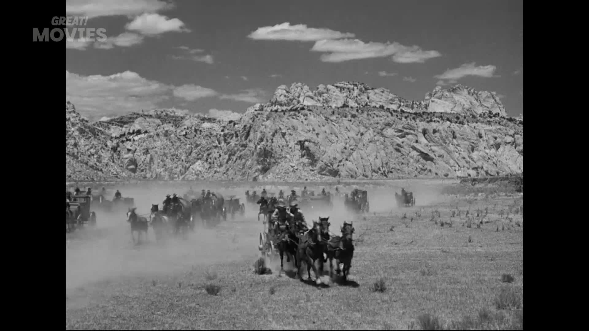A caravan of horse-drawn wagons kicks up dust as it thunders across a dry, open plain. The wagons are strung out, with a few leading the charge towards the rugged, sun-baked mountains in the distance.