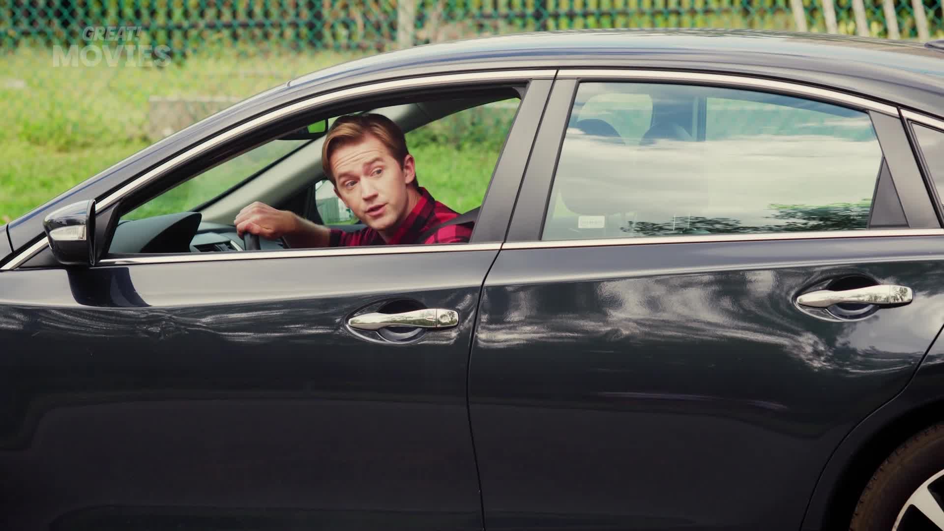 A young man in a red plaid shirt grips the steering wheel of a dark sedan. He glances over his shoulder, his expression one of mild surprise.